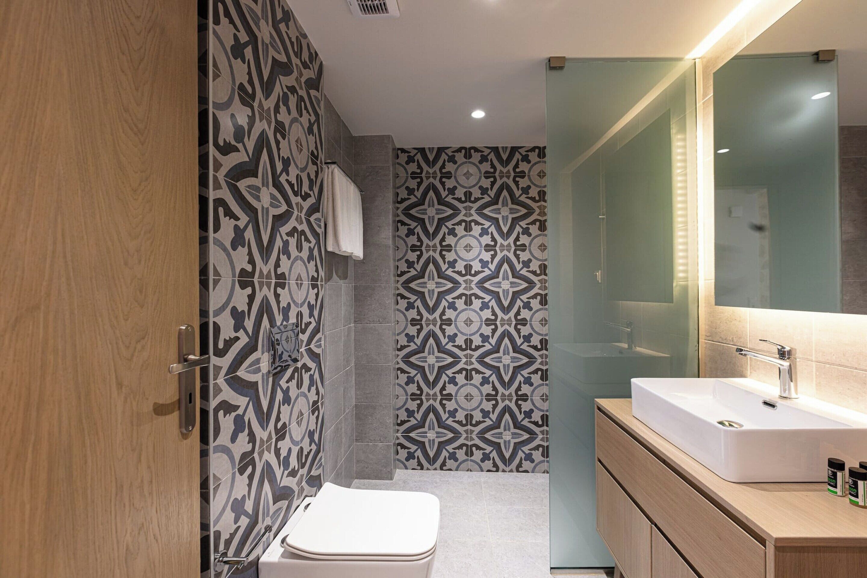 A modern bathroom with patterned tiles, a white toilet, a sink with a mirror, and wooden cabinetry.