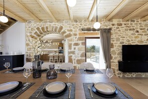 A dining area with a stone wall, wooden ceiling beams, and a TV mounted on the wall. - New Luxurious Villa With Stunning Pool,views &bbq! (Rethymno)