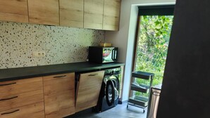 A kitchen with wooden cabinets, a countertop, a washing machine, and a view of greenery outside. - Plein Sud La Roche en Ardenne (La Roche-en-Ardenne)