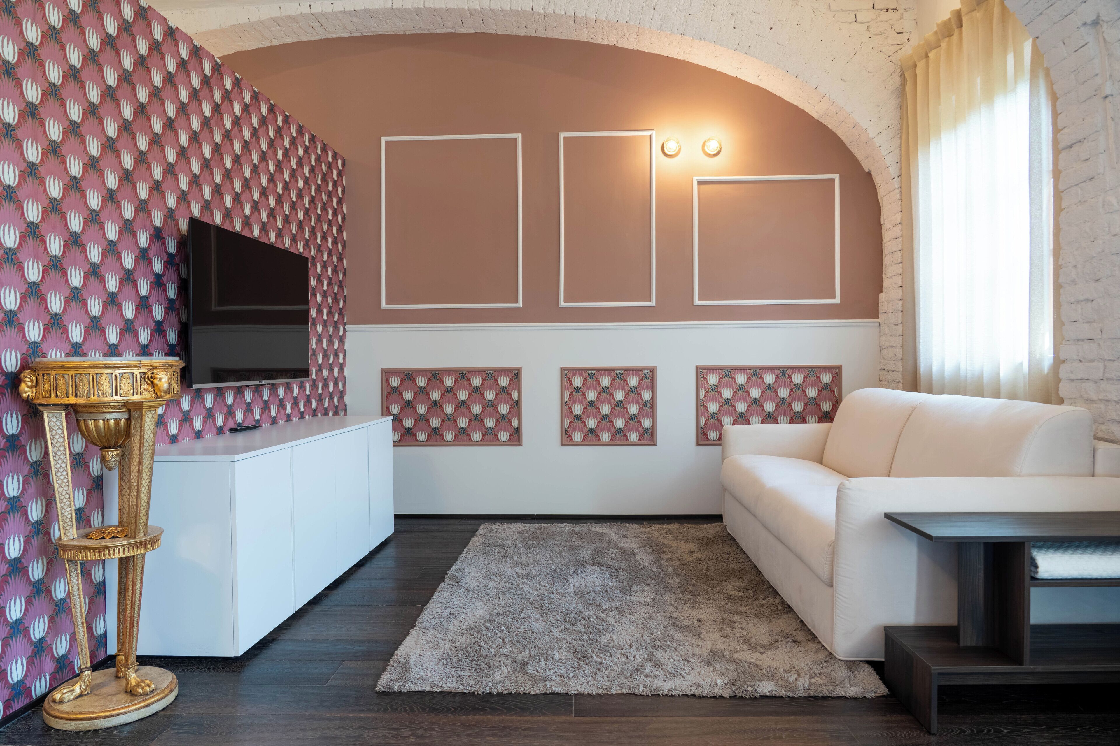 Een woonkamer met een witte bank, een televisie op een wit meubelstuk, behang met patroon en een decoratieve gouden standaard.