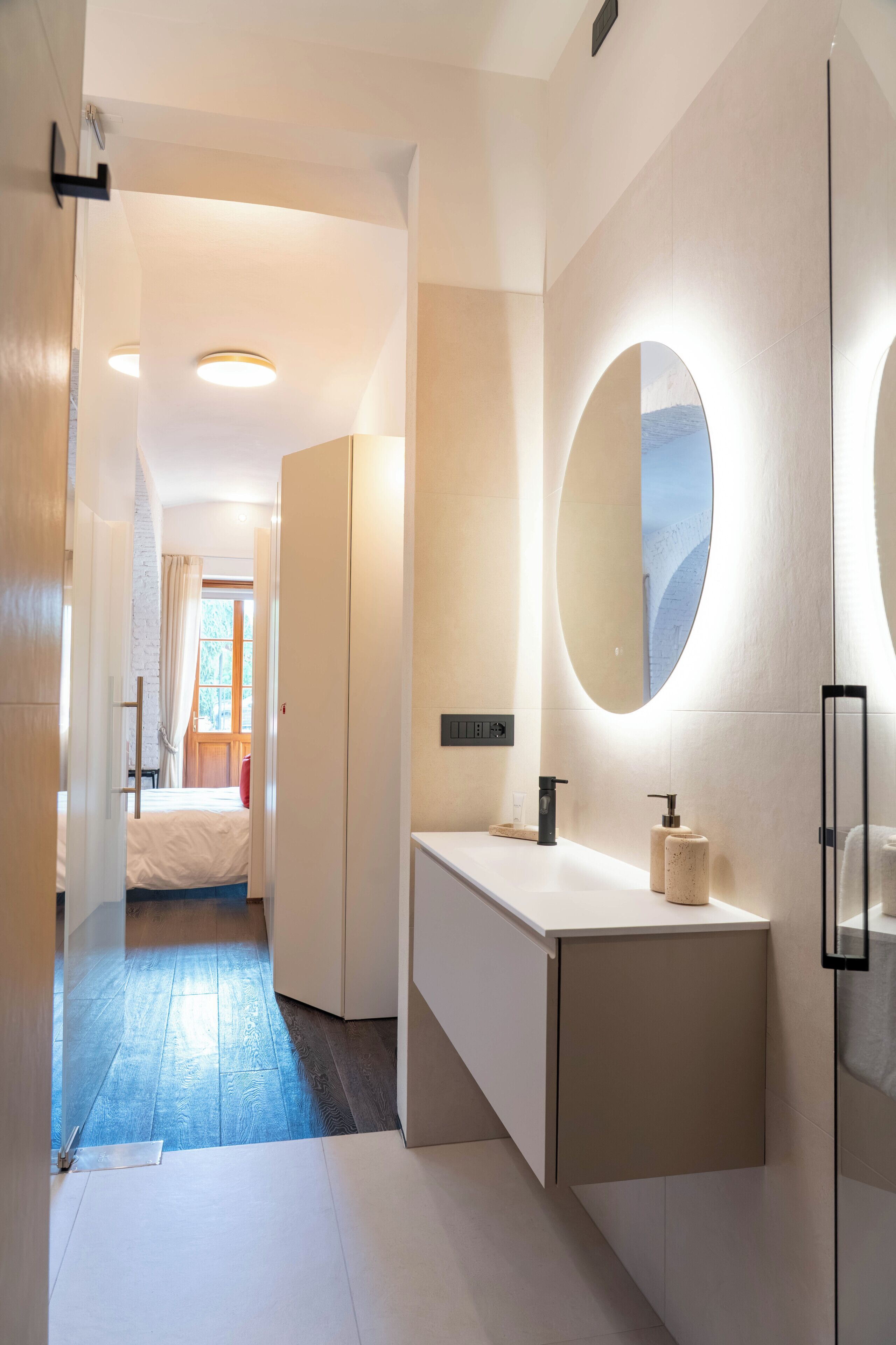 A modern bathroom with a white vanity, a large mirror, and a door leading to a bedroom.