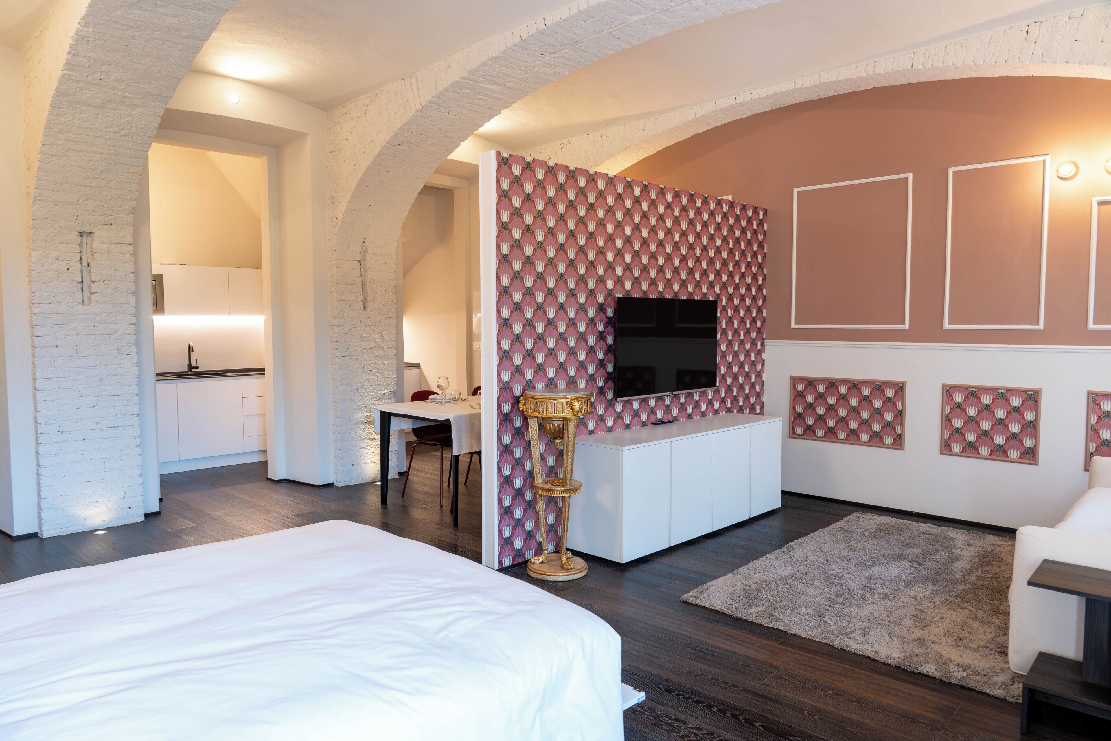 A modern hotel room with a large bed, a dining area, a kitchen, and a TV mounted on the wall.