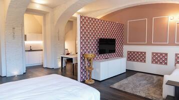 A modern hotel room with a large bed, a dining area, a kitchen, and a TV mounted on the wall.