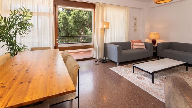 Una sala de estar moderna con una mesa de comedor de madera, sofás grises, una lámpara de pie y vistas a través de un ventanal grande.