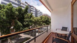 Un balcón con mesa y sillas, con vista a una calle con árboles y edificios.