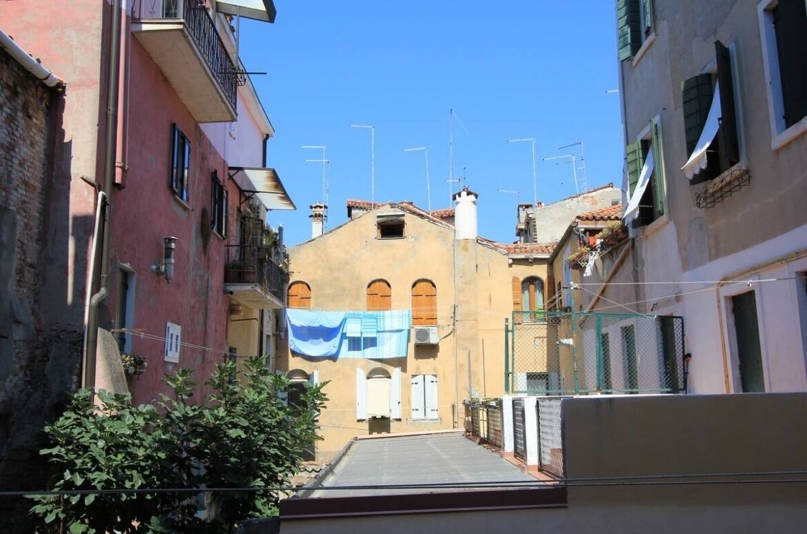 Une rue étroite, bordée de bâtiments des deux côtés, un ciel dégagé et un tissu bleu accroché à l’un des bâtiments.