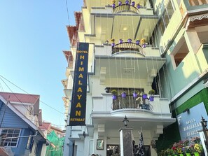 Um edifício de vários andares com varandas decoradas com vasos de plantas. É visível uma faixa com a inscrição 'HOTEL HIMALAYAN RETREAT'.
