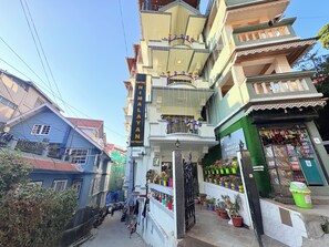Um edifício de vários andares com varandas, uma placa indicando Hotel Himalayan Retreat e uma pequena loja no nível térreo.