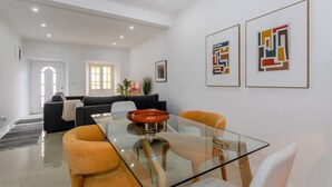 A modern living room with a glass dining table, orange chairs, a dark sofa, and abstract art on the walls.