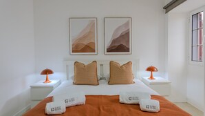 A hotel room with a bed, two pillows, two nightstands, two lamps, two framed abstract paintings, and a window.