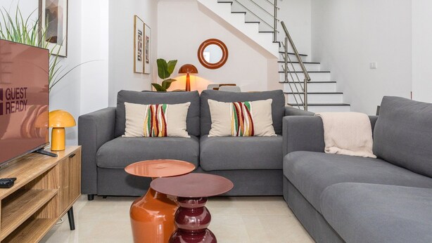 A modern living room with a grey sofa, a small wooden side table, a round mirror, and a staircase in the background.