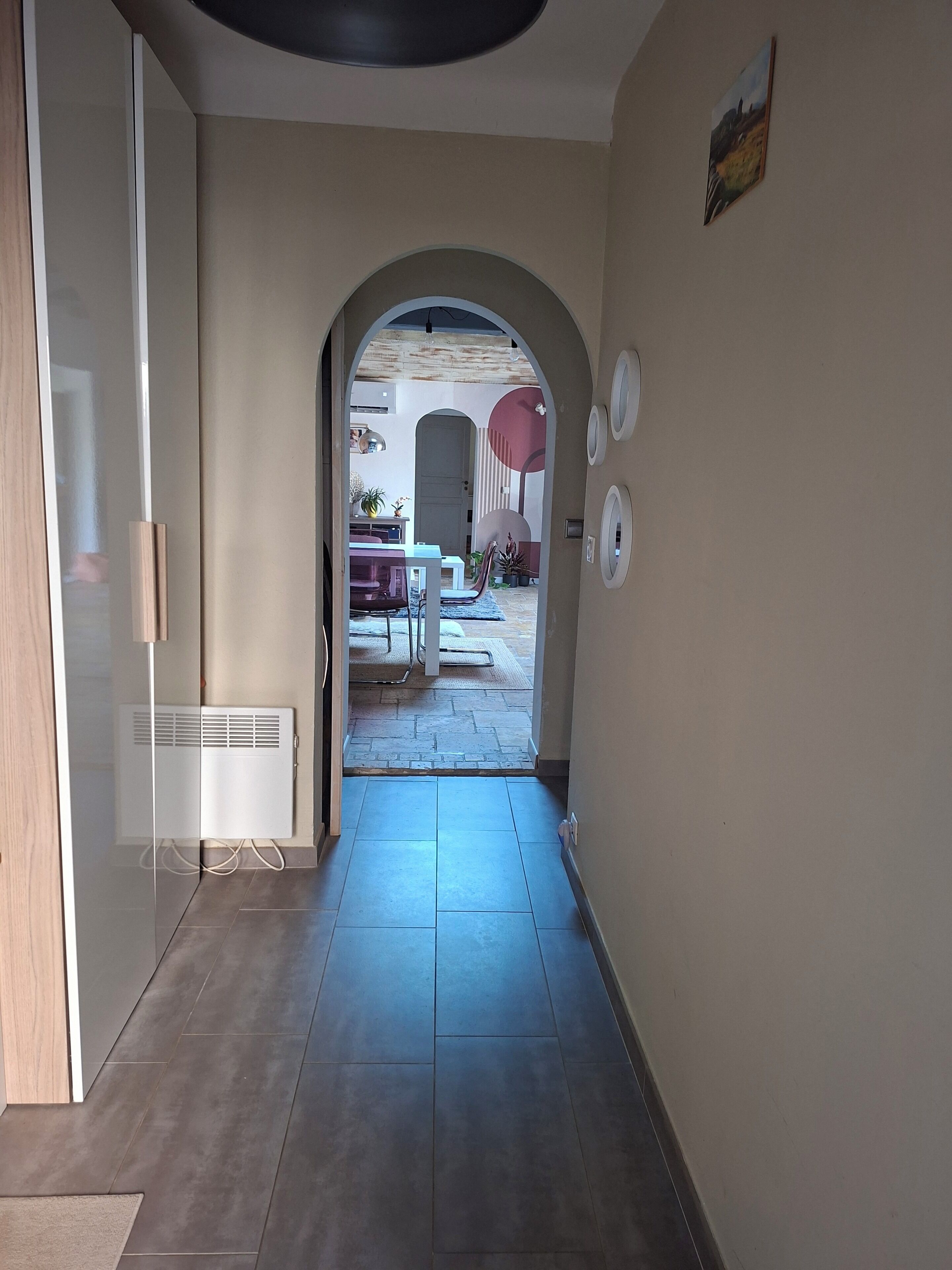 Un pasillo con suelo de baldosas, un arco de entrada a un patio exterior con mesa y sillas, y un radiador a la izquierda.