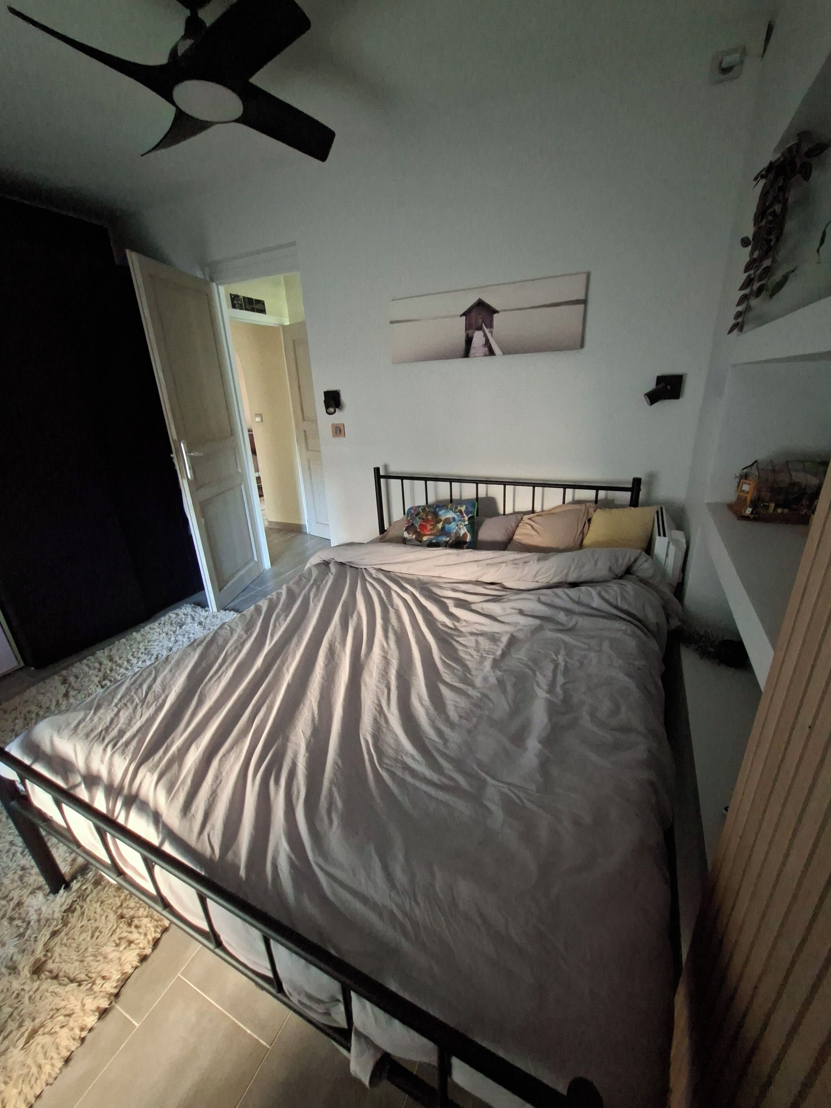 Ein Schlafzimmer mit einem Bett, einem Deckenventilator, einem an der Wand befestigten Kunstwerk und einer Tür zu einem anderen Raum.