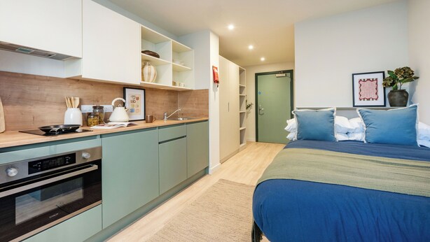 Habitación de hotel compacta con kitchenette, cama con ropa de cama azul, mesita de noche y un cuadro enmarcado en la pared.