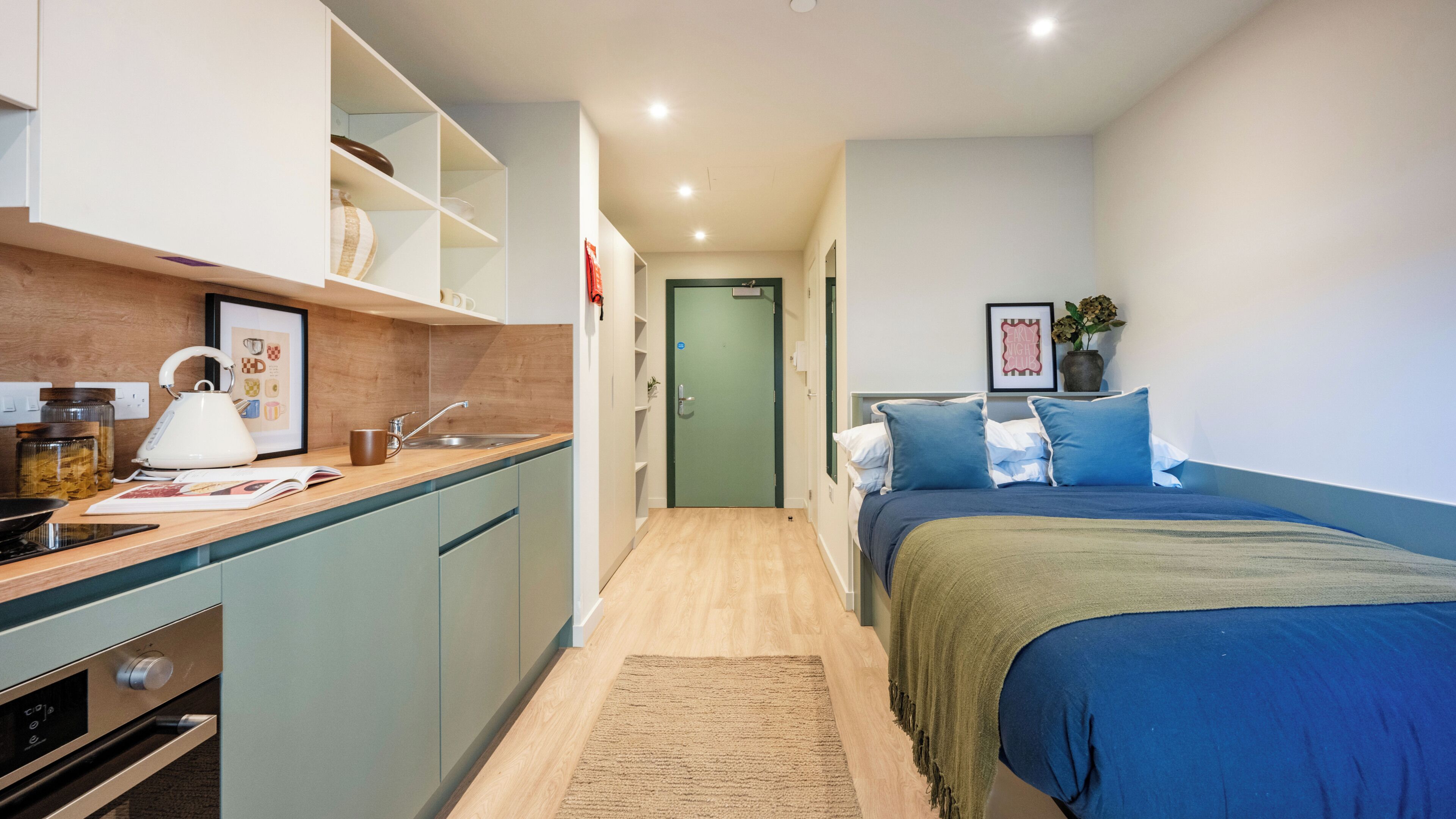A compact hotel room with a kitchenette, a bed with blue bedding, a green door, and a framed picture on the wall.