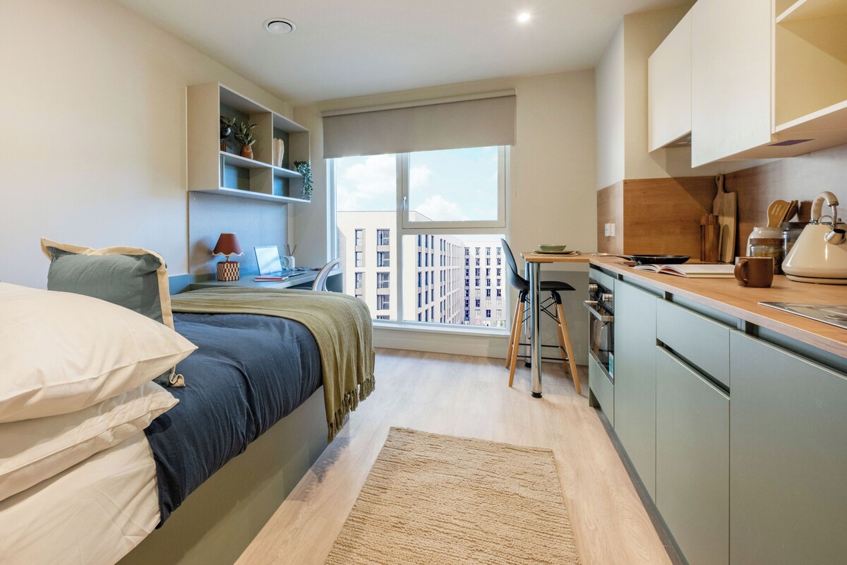 Quarto moderno com cama, mesa de cabeceira e vista para a paisagem urbana através de uma grande janela.