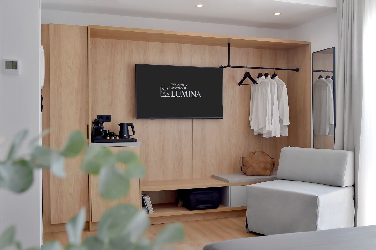 A modern hotel room with a flat-screen TV mounted on a wooden wall, a seating area with a sofa, a coat rack with hanging clothes, and a small table with a coffee maker.
