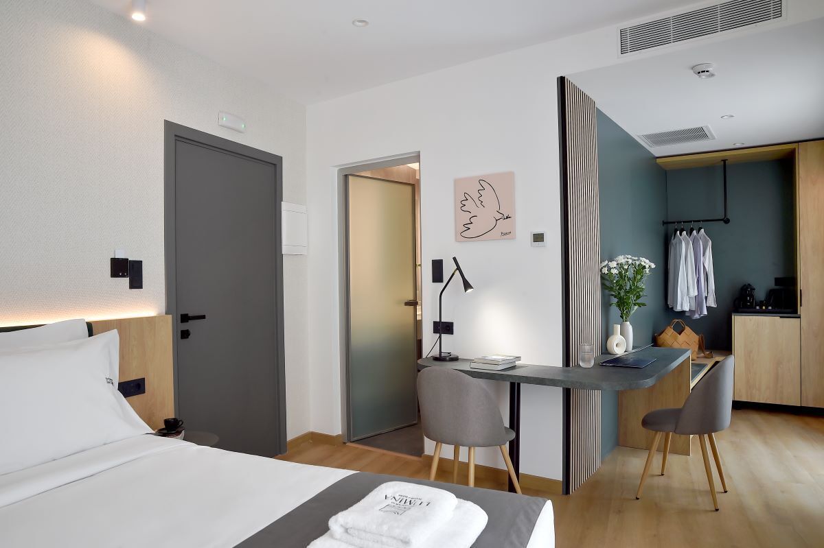 A modern hotel room with a bed, desk, and kitchenette.