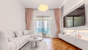 Un moderno salón con un sofá blanco, una mesa redonda de centro, un televisor de pantalla plana y vistas de la ciudad a través de amplias ventanas.