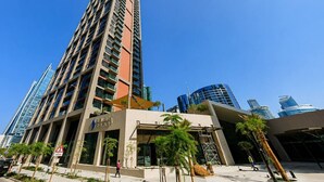 A modern high-rise building with a prominent entrance, surrounded by other tall buildings, palm trees, and a clear sky. - HAVN - Studio in Peninsula 2, Business Bay (Dubai)