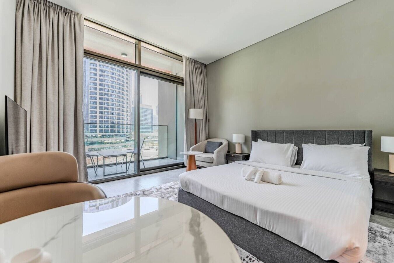Ein Hotelzimmer mit einem großen Bett, einem Sessel, einem Tisch und Blick auf eine Stadtlandschaft durch große Fenster.