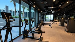 A gym with exercise equipment, large windows, and a view of palm trees and buildings. - HAVN - Studio in Peninsula 2, Business Bay (Dubai)