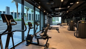 A gym with exercise equipment, large windows, and a view of palm trees and buildings. - HAVN - Modern Studio in Peninsula, Business Bay (Dubai)