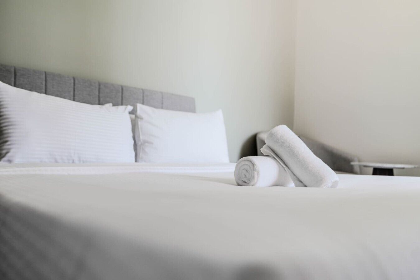 A neatly made bed with white linens and pillows, a rolled towel, and a small bedside table.