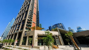 A modern high-rise building with a prominent entrance, surrounded by other tall buildings, palm trees, and a clear sky. - HAVN - Modern Studio in Peninsula, Business Bay (Dubai)