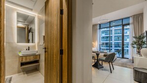 Habitación de hotel moderna con puerta de madera, baño con lavamanos y espejo, zona de comedor con mesa y sillas, y ventanas amplias con vistas a la ciudad.