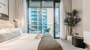 Una habitación de hotel moderna con una cama grande, un balcón con vistas a rascacielos, una planta en maceta y una silla.