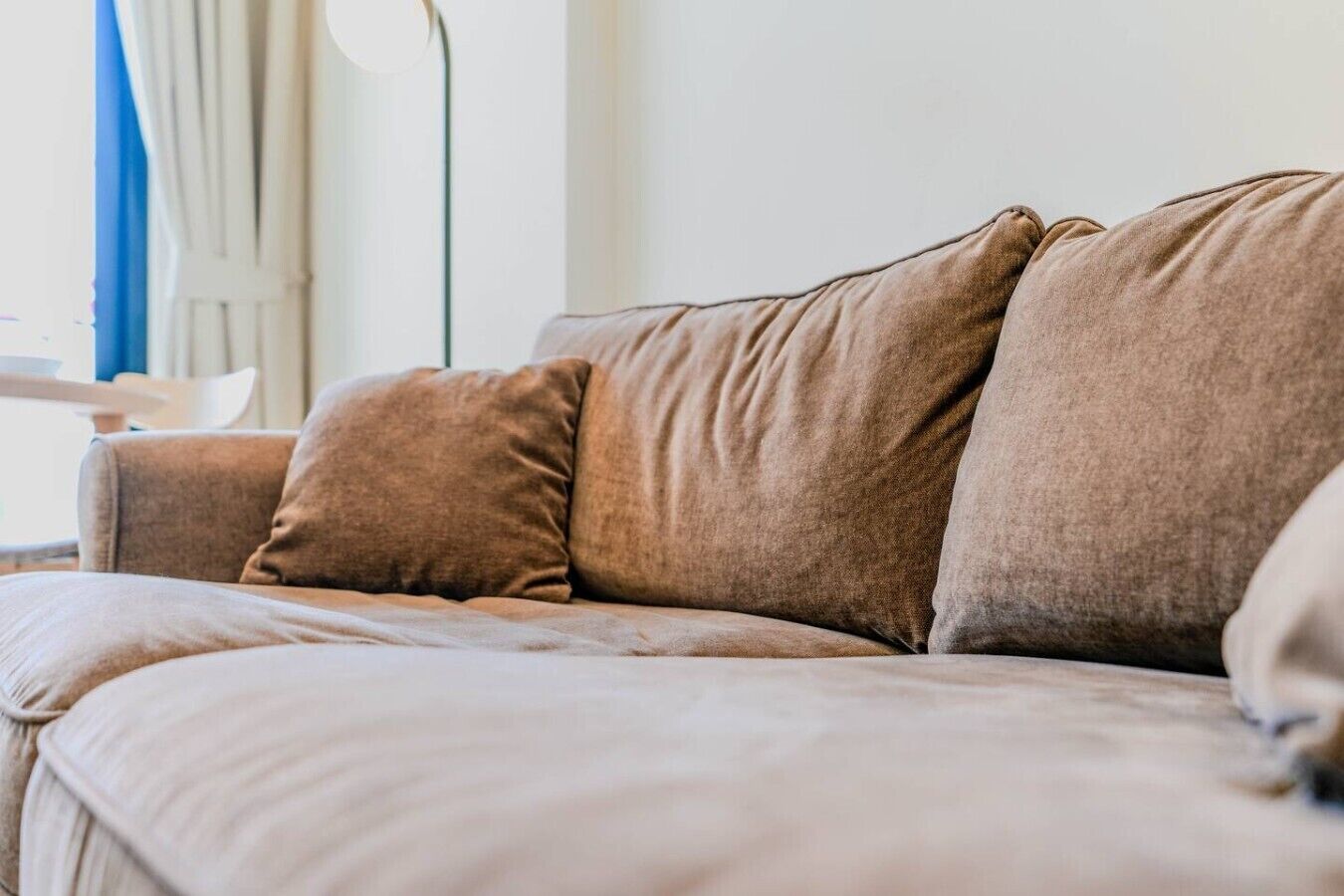 A beige sofa with multiple cushions in a room with a white wall and a blue curtain.