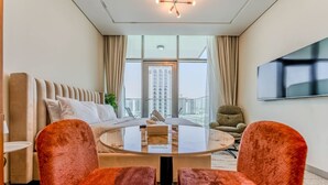 A modern hotel room with a sofa, two armchairs, a round table, a TV, and a large window with a city view. - HAVN - Studio in PG One, Al Furjan (Dubai)