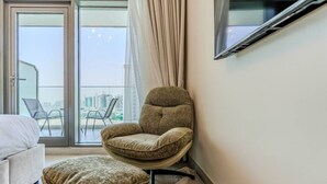 A modern hotel room with a large sliding glass door leading to a balcony, a comfortable armchair, a matching ottoman, and a flat-screen TV mounted on the wall. - HAVN - Studio in PG One, Al Furjan (Dubai)