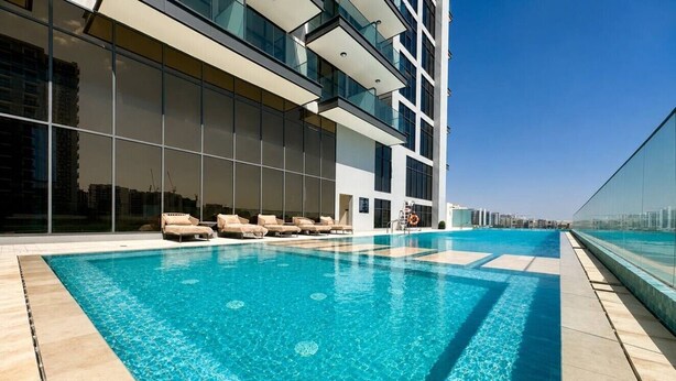 A modern building with a large outdoor swimming pool, lounge chairs, and a glass railing. - HAVN - Studio in PG One, Al Furjan (Dubai)