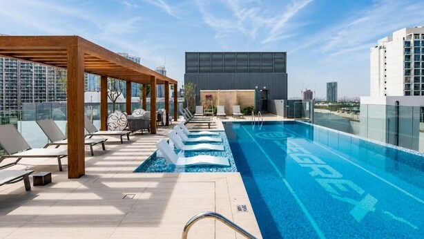 A rooftop pool with lounge chairs, a pergola, and a modern building in the background. - HAVN - Studio in Stonehenge Residences, JVC (Dubai)