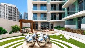 A modern building with a courtyard featuring a central sculpture, a seating area, and a view of the surrounding high-rise buildings. - HAVN - Studio in Stonehenge Residences, JVC (Dubai)