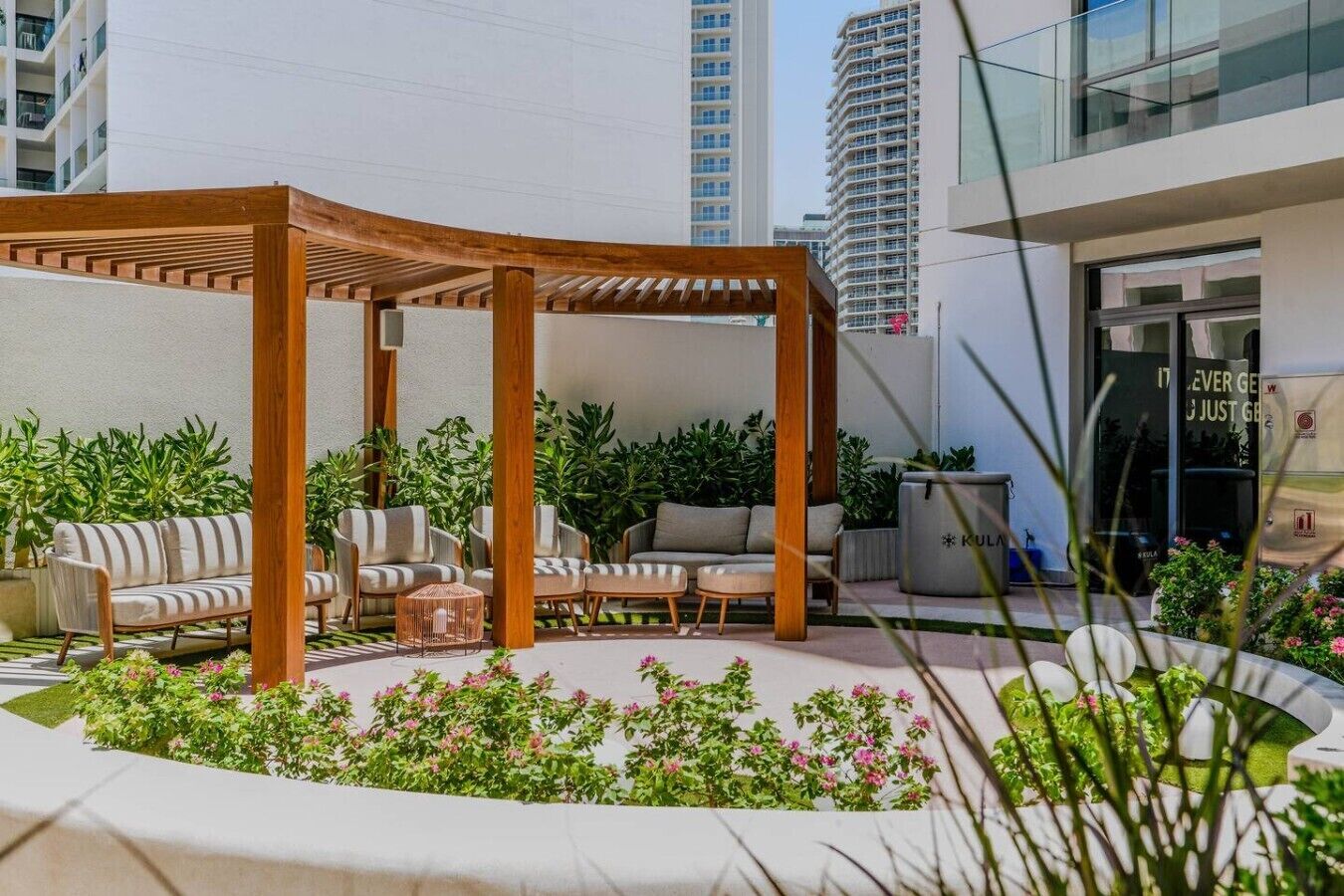 A covered outdoor seating area with sofas and chairs, surrounded by greenery and flowers.