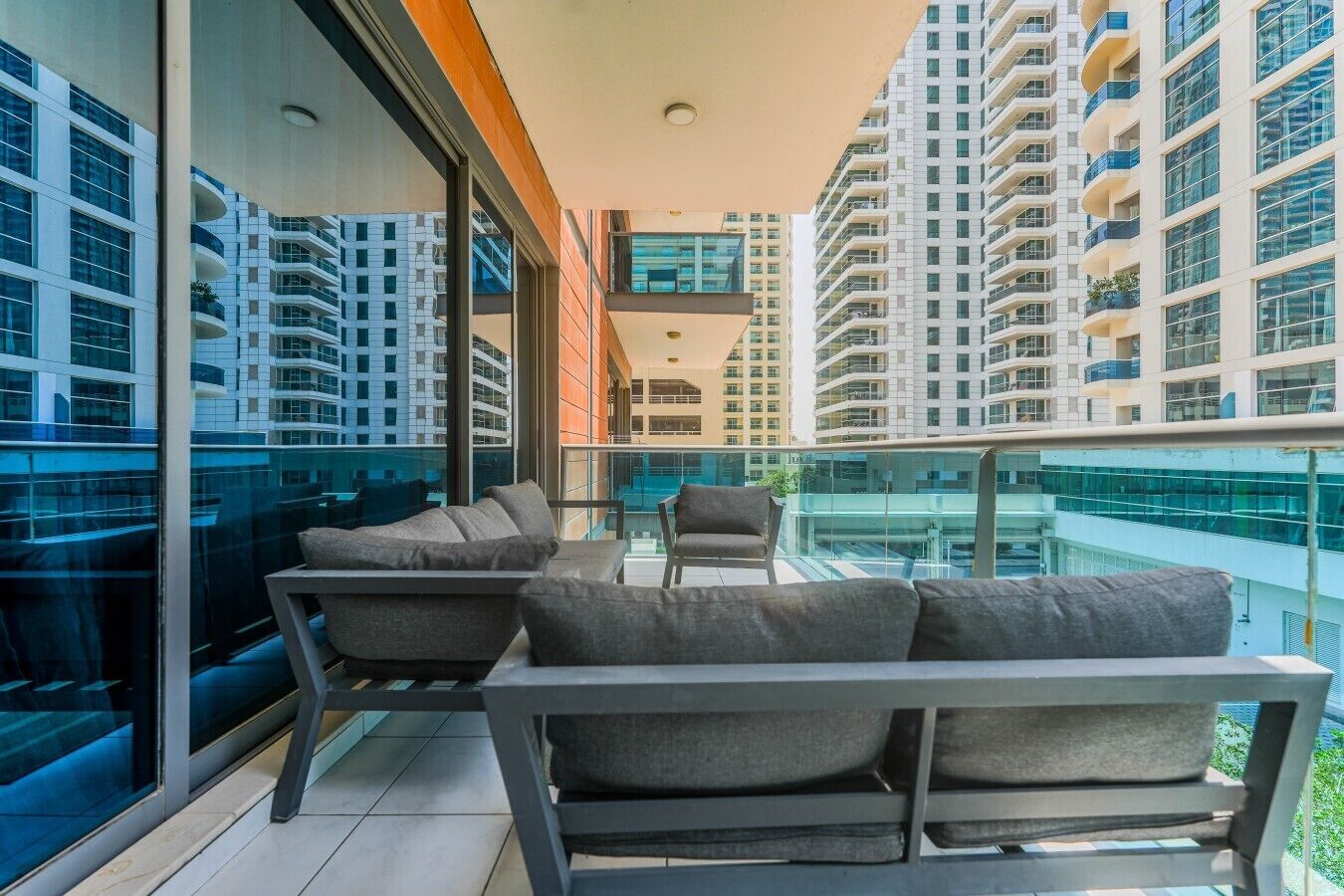 Een balkon met een bank en stoelen, grote ramen en uitzicht op hoogbouw.