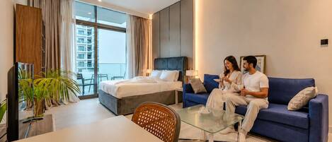A hotel room with a bed, a sofa, a glass coffee table, a TV, and a balcony with a view of buildings.