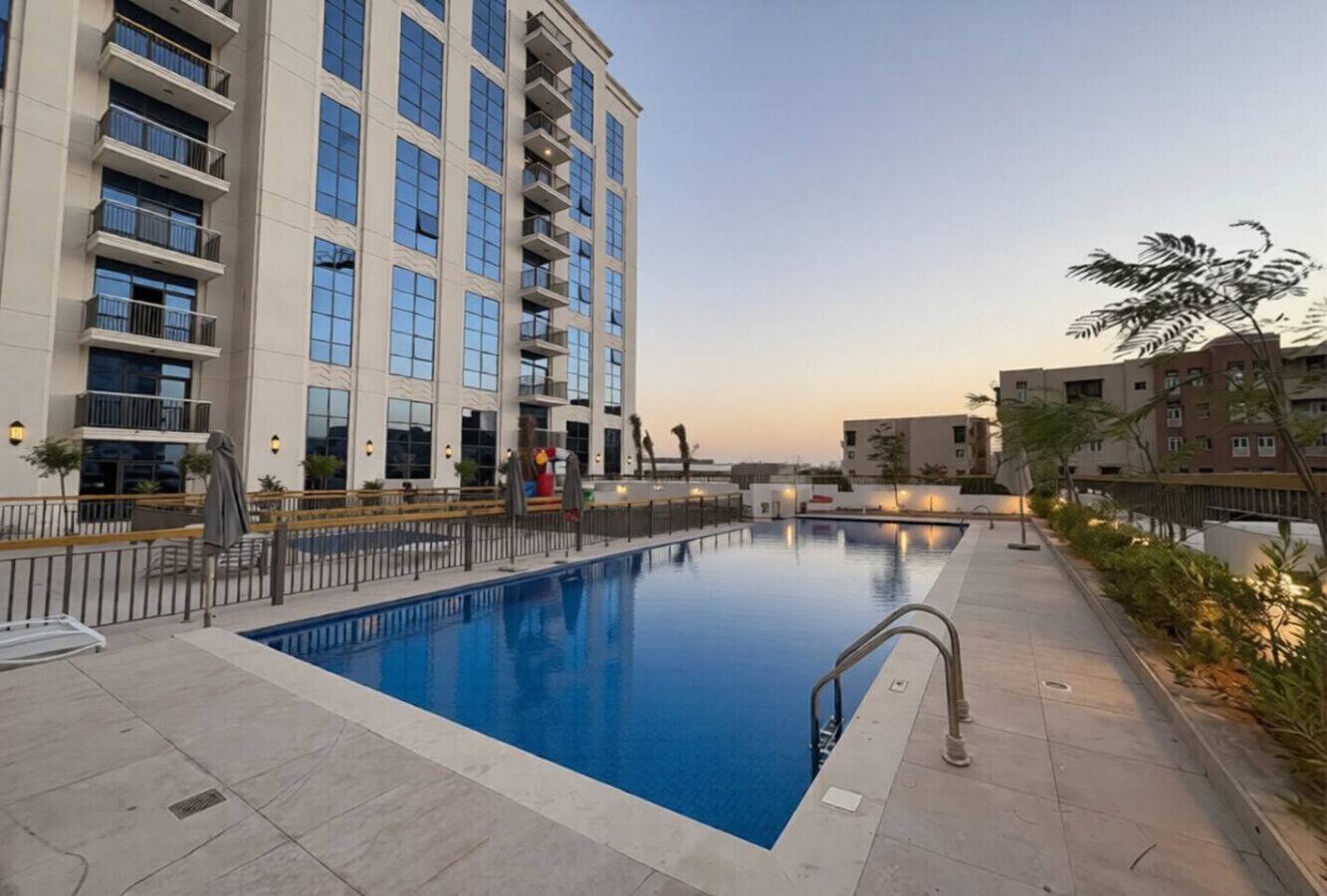 A swimming pool with a metal ladder, surrounded by a fence, and adjacent to a multi-story building with balconies.