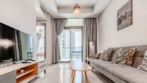 A modern living room with a flat-screen TV, a sofa with patterned cushions, a coffee table, and a large window with curtains. - HAVN - 2 Bedroom in Aykon Tower, Business bay (Dubai)