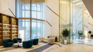 A spacious lobby with a modern design, featuring a seating area with sofas and armchairs, a large window with natural light, and a decorative wall with shelves. - HAVN - 2 Bedroom in Aykon Tower, Business bay (Dubai)