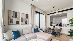 Ein modernes Wohnzimmer mit einer Eckcouch, einem runden Couchtisch, abstrakter Wandkunst, einem Essbereich mit Tisch und Stühlen sowie einer Küche im Hintergrund.