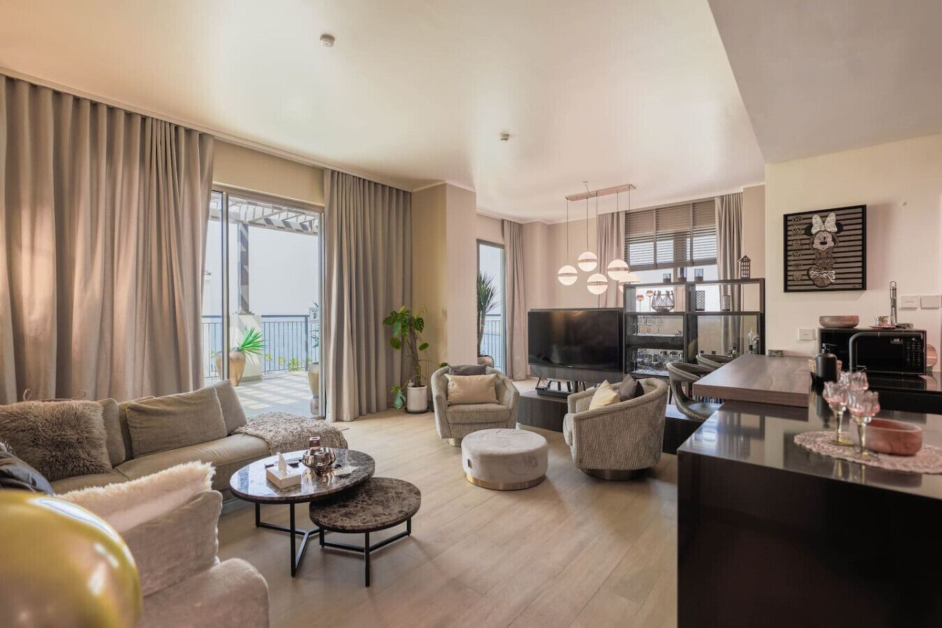 A spacious living room with a large flat-screen TV, comfortable seating, a dining area, and a balcony with curtains.
