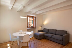 A modern living room with a grey sofa, a white dining table with chairs, a pendant light, and a window with a view of a building. - Paola's House - In the Heart of Bardolino (Bardolino)