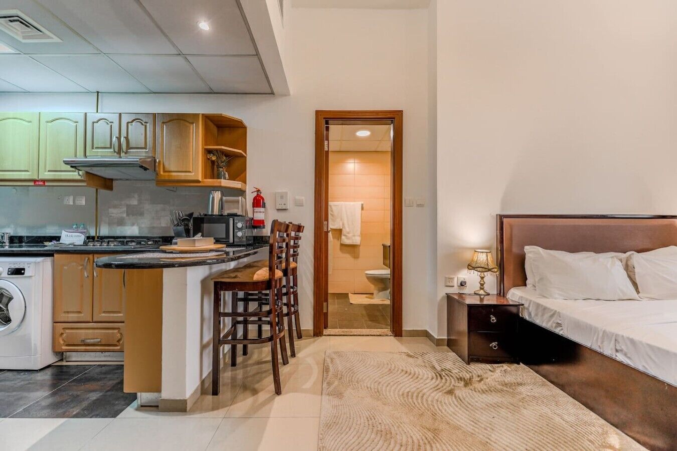 Ein kleines Hotelzimmer mit einer Küchenzeile, einem Bett, einem Nachttisch und einem durch eine geöffnete Tür einsehbaren Badezimmer.