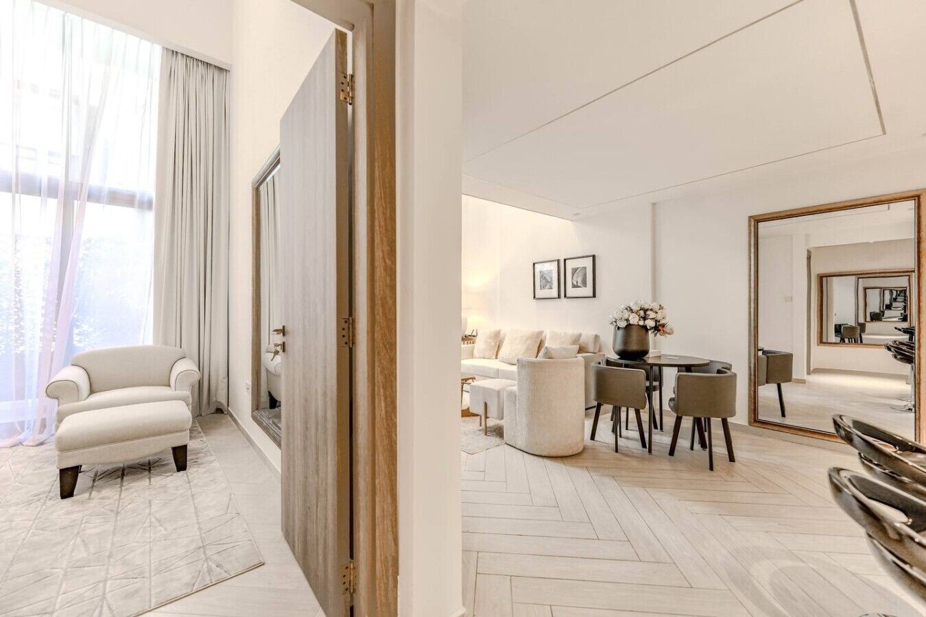 A modern hotel room with a sofa, a dining table with chairs, a mirror, and a view through a door.