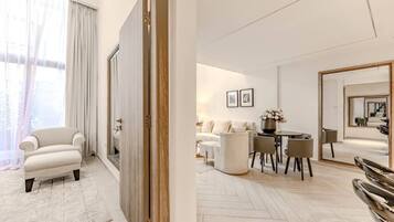 A modern hotel room with a sofa, a dining table with chairs, a mirror, and a view through a door.