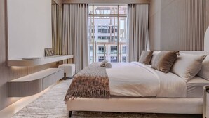 Une chambre d’hôtel moderne, dotée d’un grand lit, d’une table de chevet, d’une banquette intégrée et offrant une vue sur le paysage urbain grâce à de grandes fenêtres du sol au plafond.